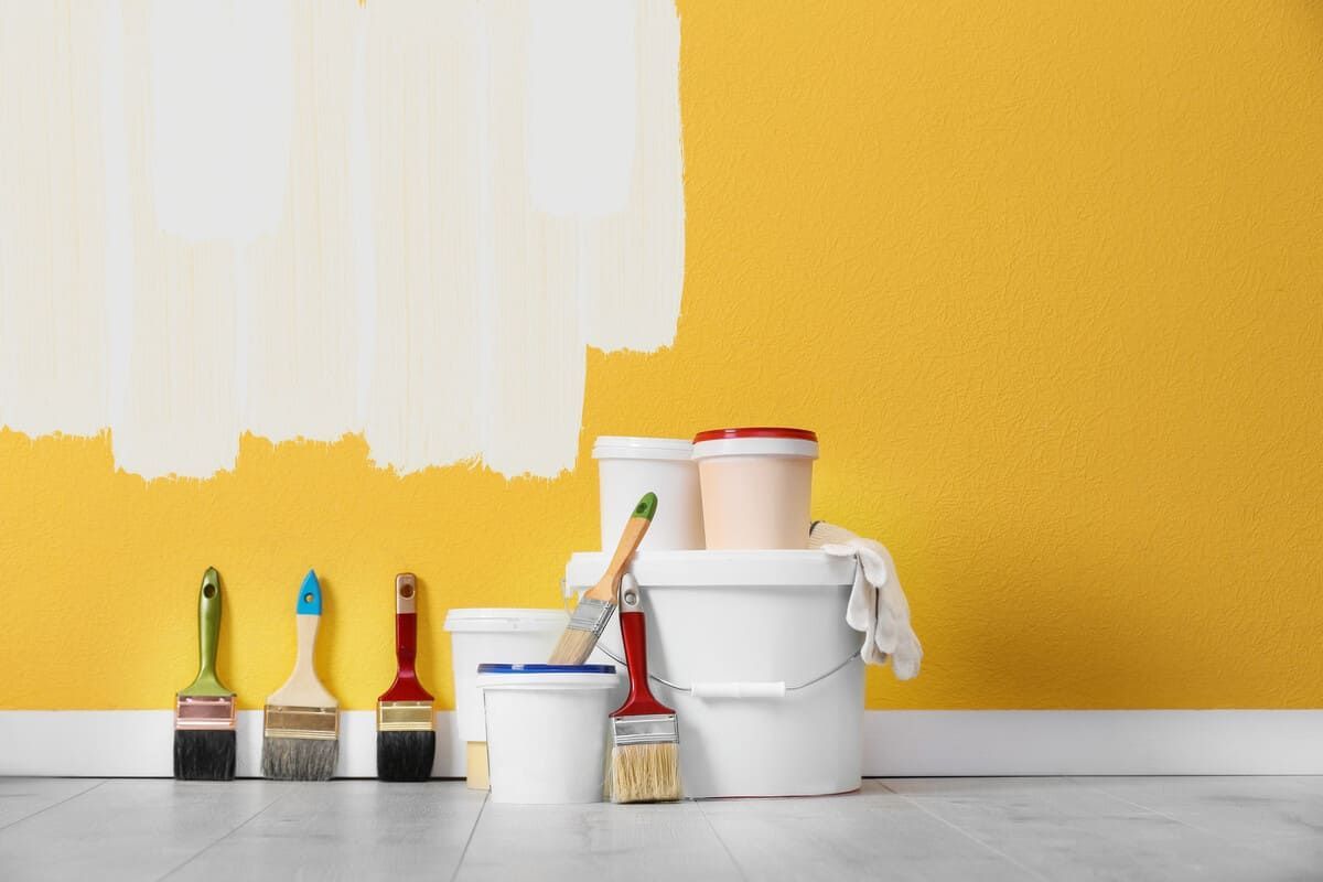 Materiales de pintura y una muestra de pintura blanca sobre una pared amarilla. Pinceles, cubos y un paño sobre un suelo de madera.