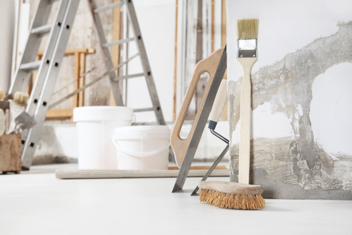 Herramientas para pintar y renovar: una escalera, baldes, paleta, brocha y rodillo contra una pared.