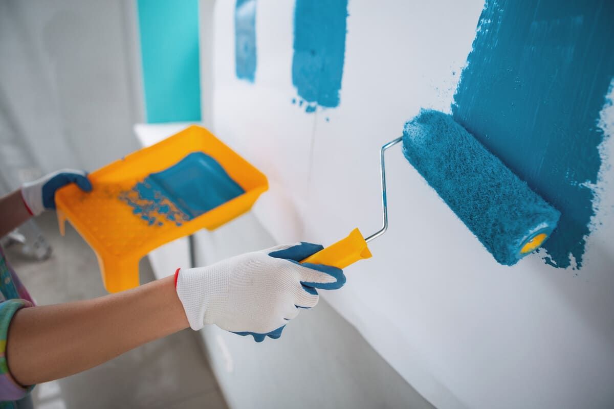 Persona pintando una pared blanca de azul con un rodillo. Lleva guantes y una bandeja de pintura amarilla.
