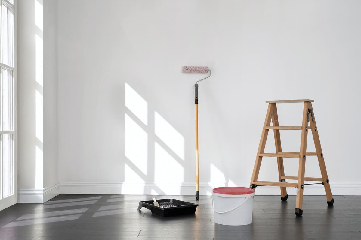 Una habitación preparada para pintar con rodillo, cubo de pintura, escalera y bandeja. La luz del sol entra a raudales por una ventana.