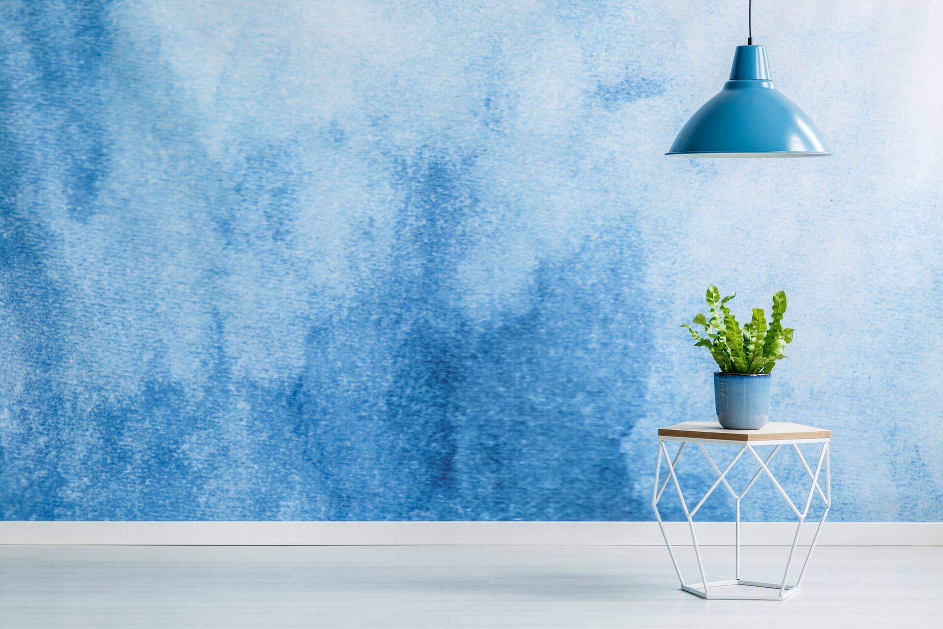 Una pared azul estilo acuarela con una lámpara colgante verde azulado, una planta sobre una mesa pequeña y un piso blanco.
