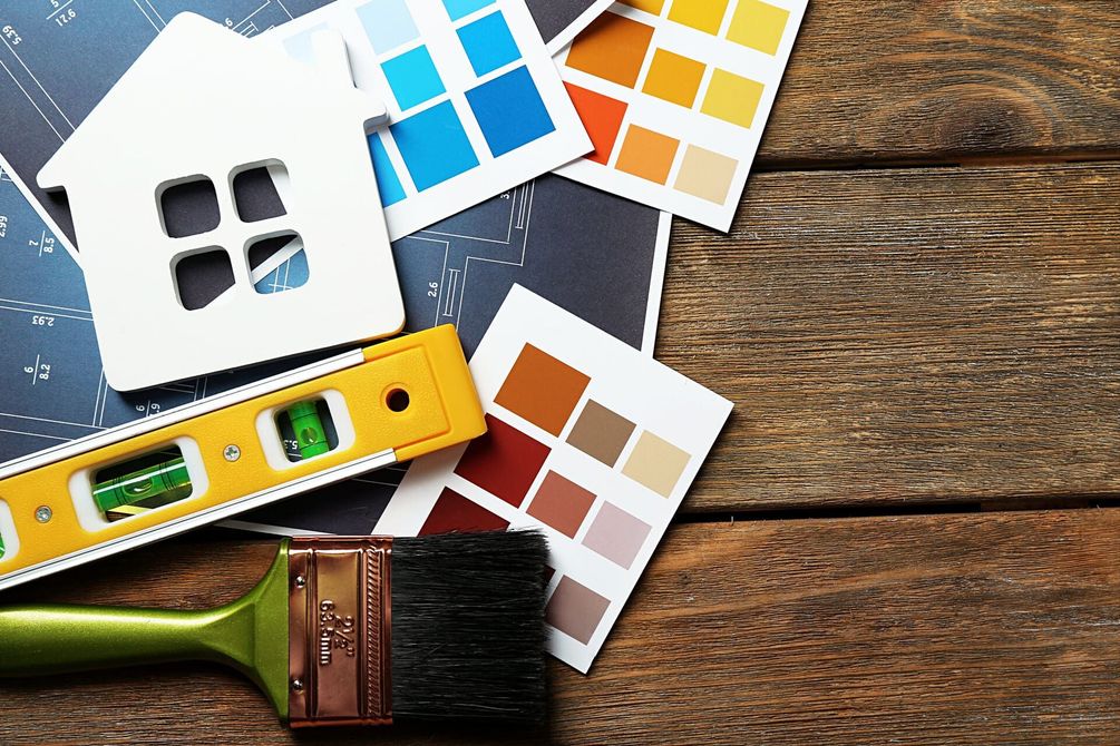 Recorte con forma de casa, muestras de pintura y nivel sobre tablones de madera, sugiriendo renovación y diseño del hogar.