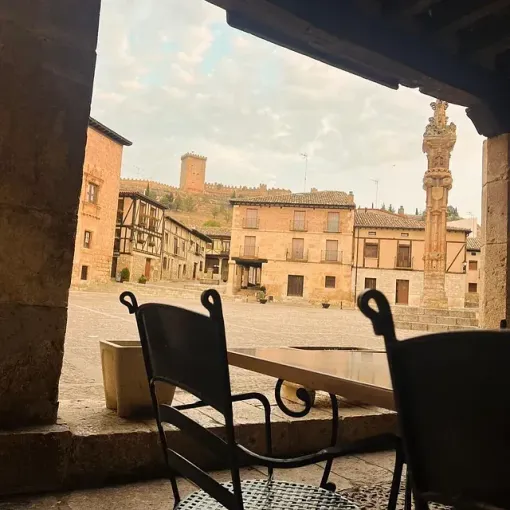 Las sillas y la mesa del café dan a una soleada plaza histórica europea con edificios antiguos y una alta torre de piedra.