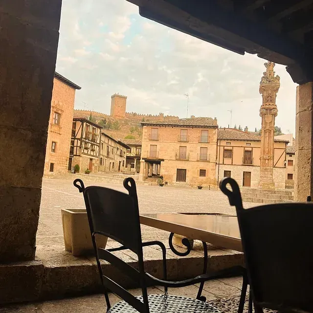 Las sillas y la mesa del café dan a una soleada plaza histórica europea con edificios antiguos y una alta torre de piedra.
