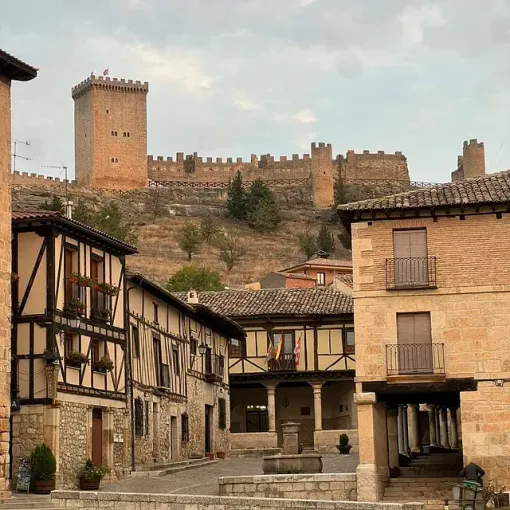 Pueblo histórico de piedra con casas de entramado de madera frente a un castillo en la cima de una colina bajo un cielo nublado.