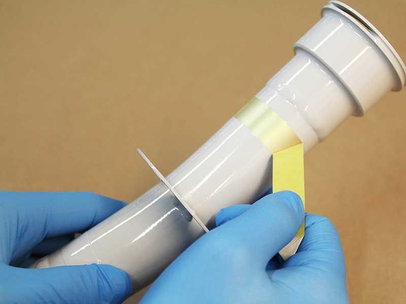 Hände in blauen Handschuhen halten ein weißes PVC-Rohr mit gelbem Klebeband. 