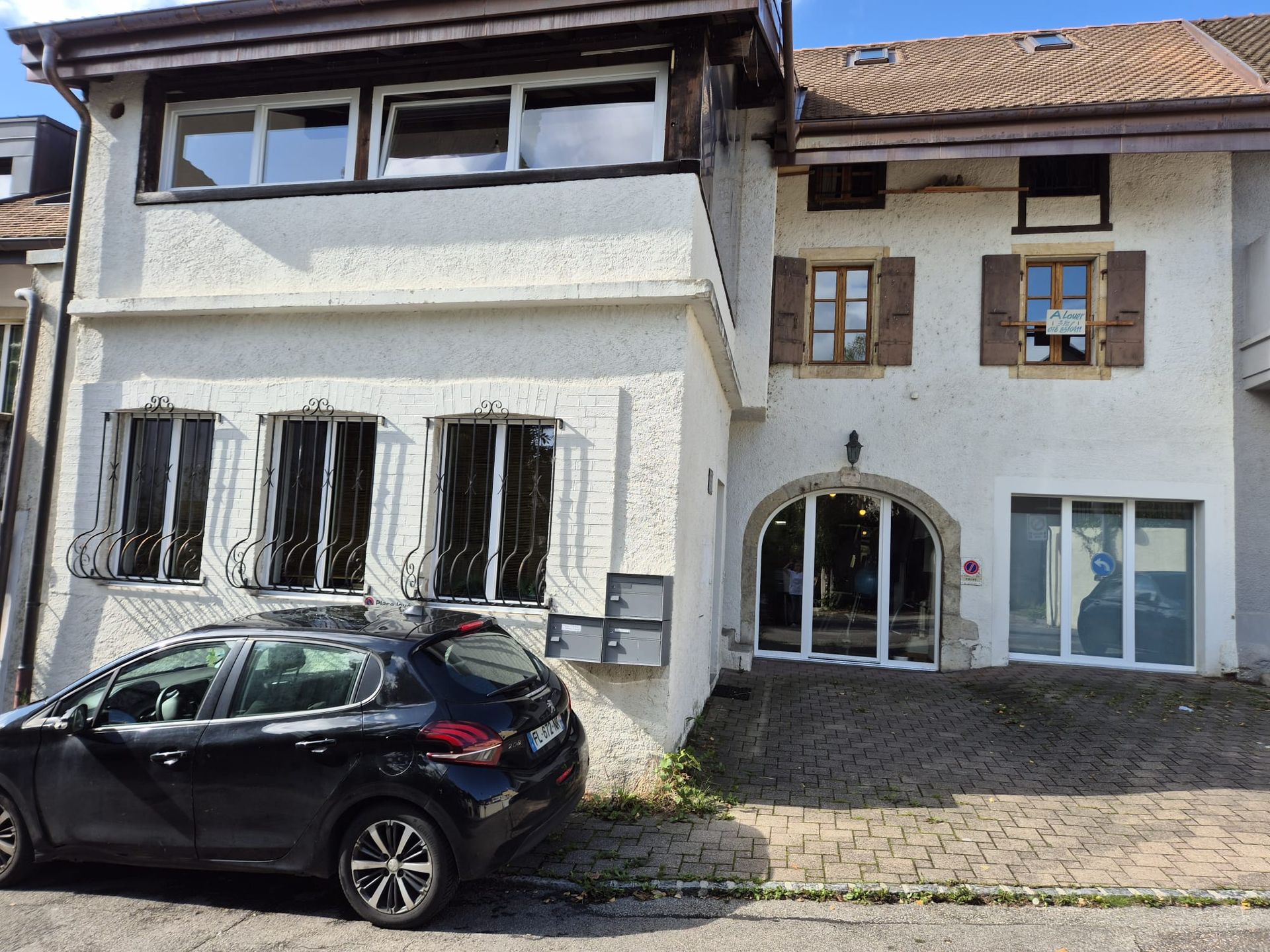 Bâtiment en stuc blanc avec fenêtres, volets et porte cintrée. Une voiture est garée devant.