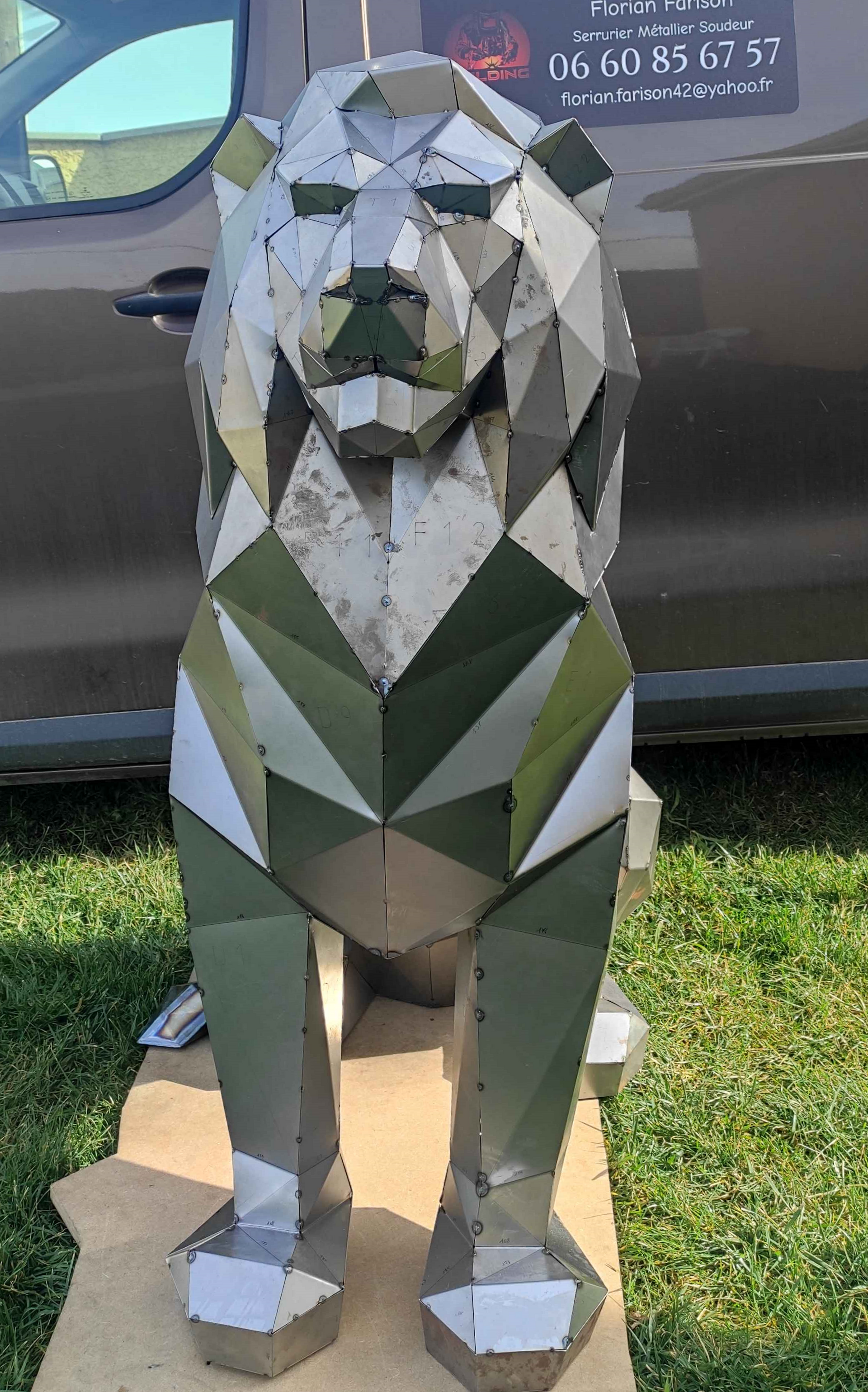 Une sculpture de lion en métal, géométrique et low-poly, aux nuances d'olive, de gris et de blanc, posée sur un socle en pierre claire à l'extérieur.