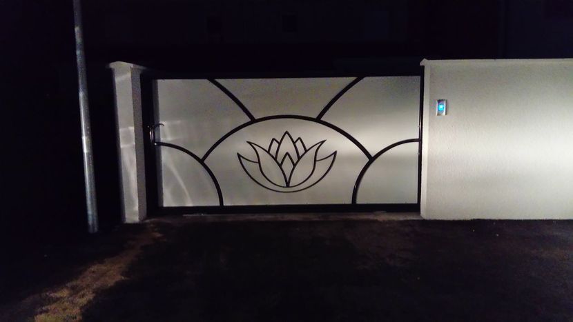 Une porte métallique la nuit, ornée d'un motif central de fleur de lotus cerné de noir sur un panneau clair.