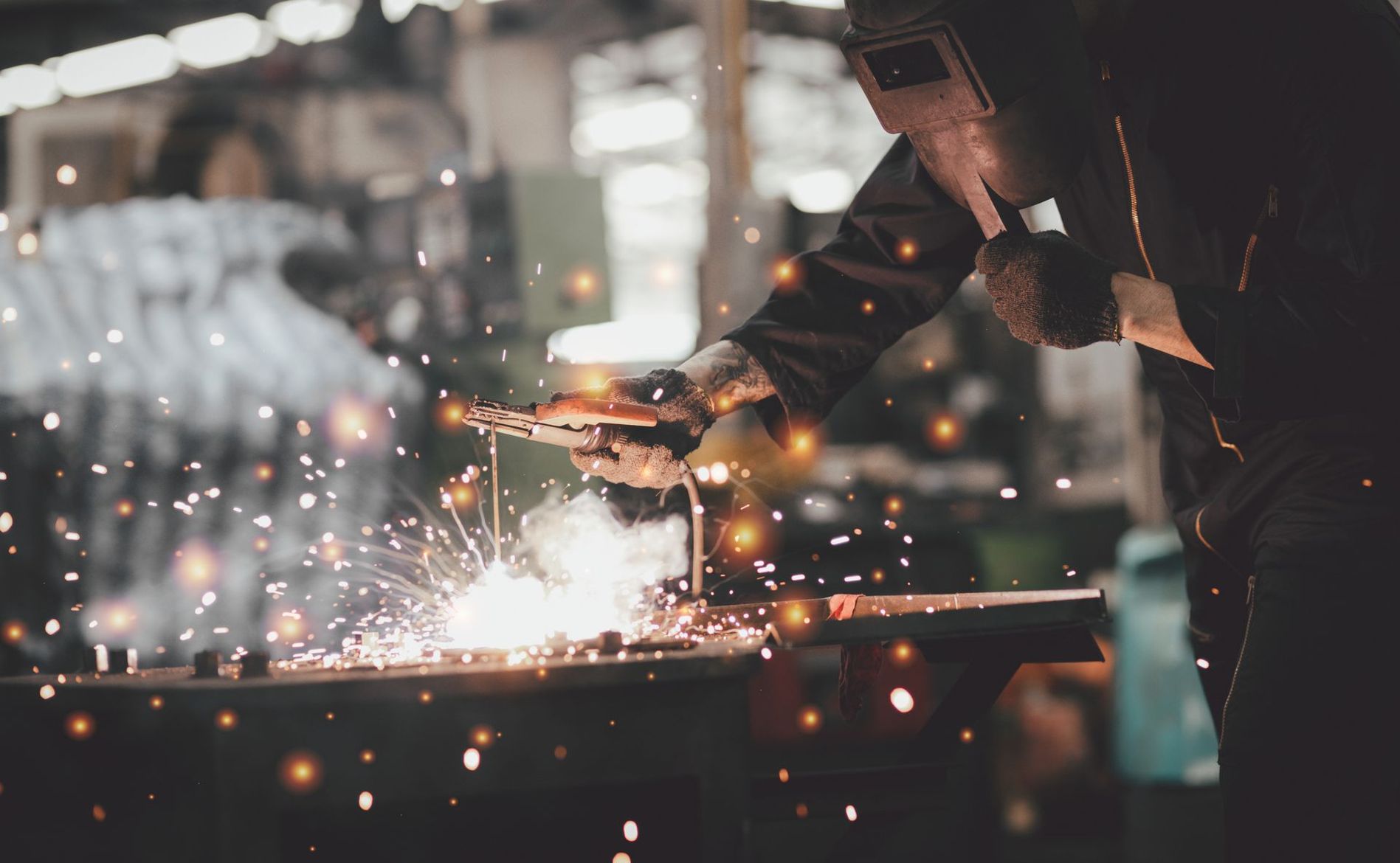 Un soudeur en tenue de protection travaille sur du métal dans une usine, entouré d'étincelles vives jaillissant du point de contact.