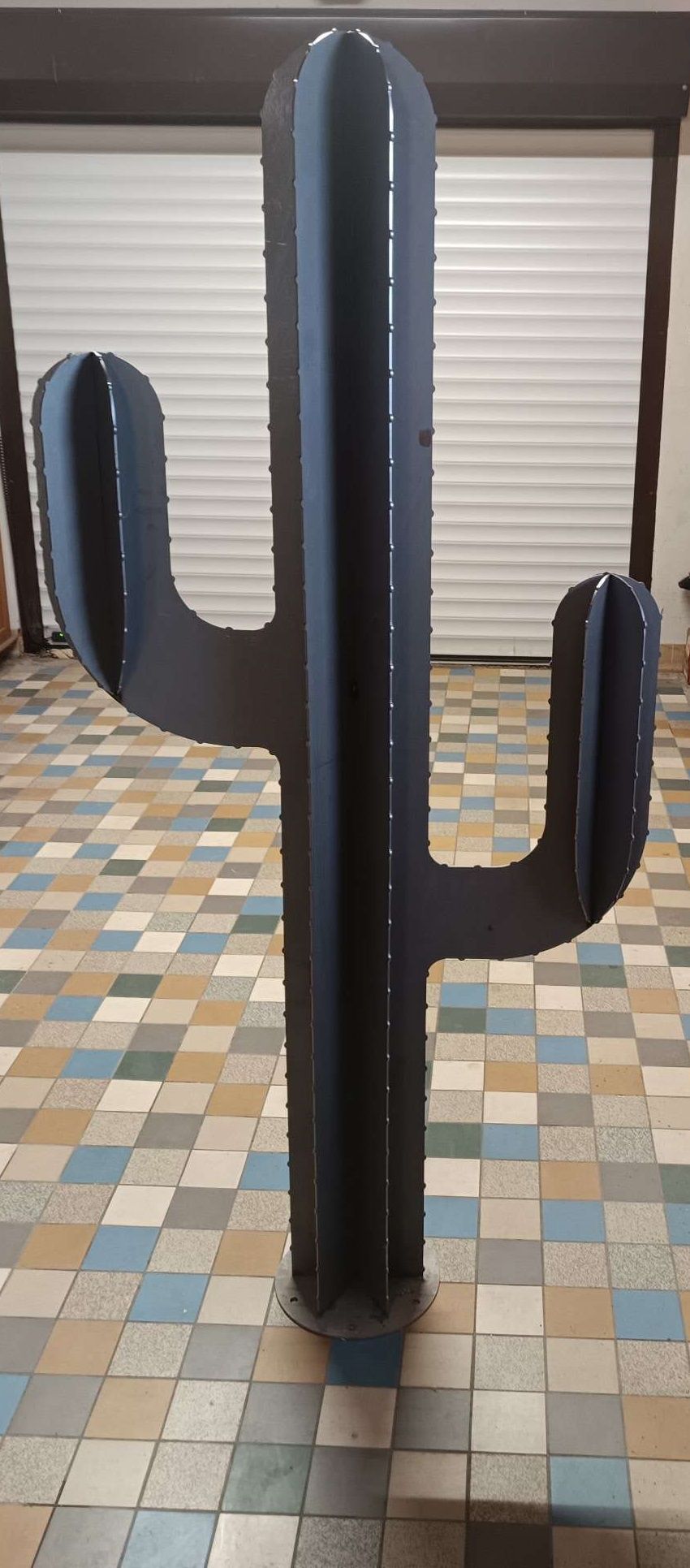 Une grande sculpture métallique stylisée, sombre et de forme allongée, représentant un cactus saguaro, posée sur un sol carrelé dans un garage.