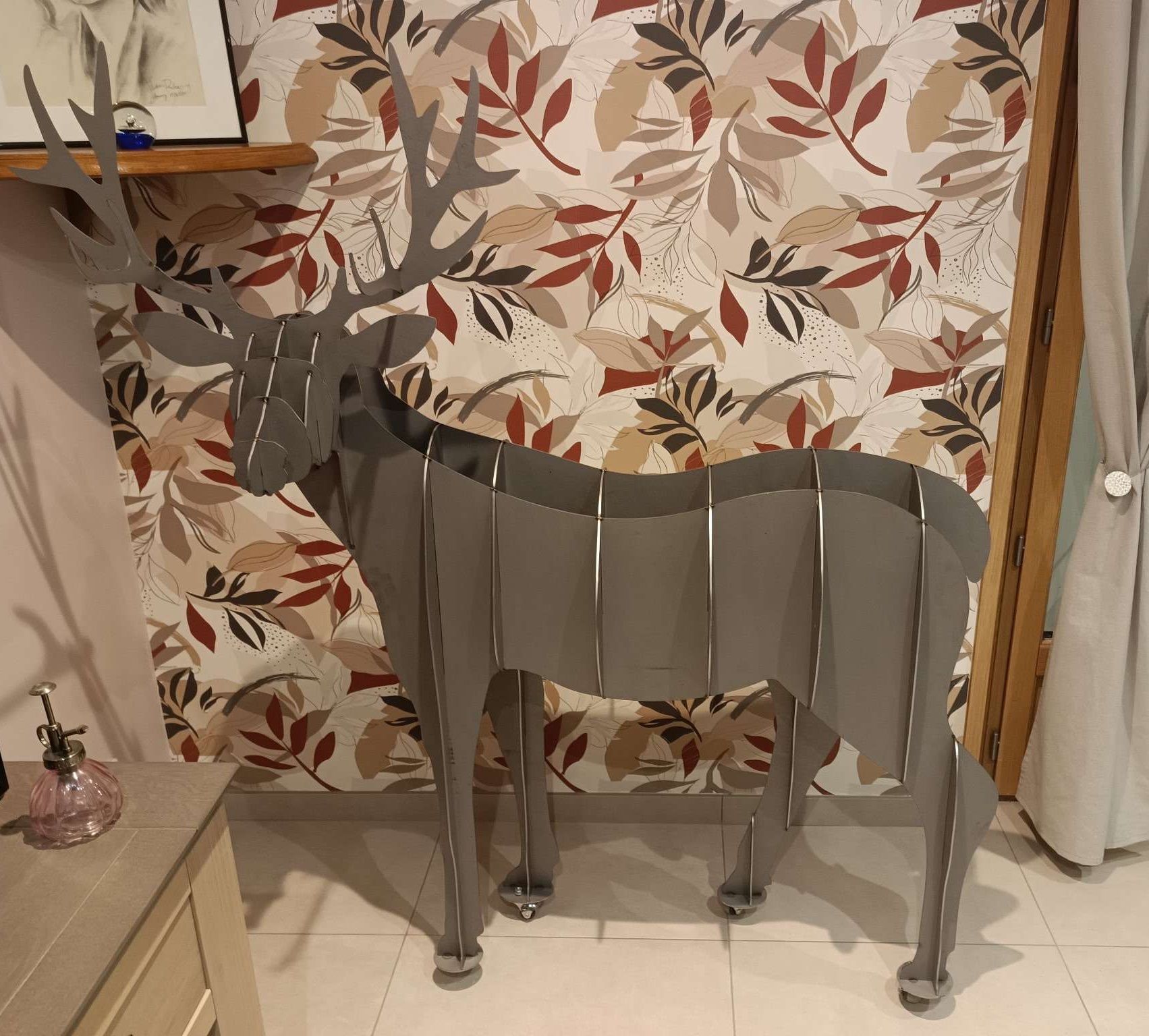Une grande sculpture grise en kit représentant un cerf, composée de planches à fentes entrecroisées, se dresse dans une pièce tapissée de papier peint à motifs.