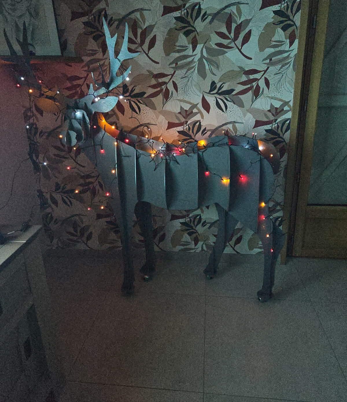 Une sculpture de cerf en carton gris aux formes géométriques, placée à l'intérieur et décorée de guirlandes lumineuses colorées.