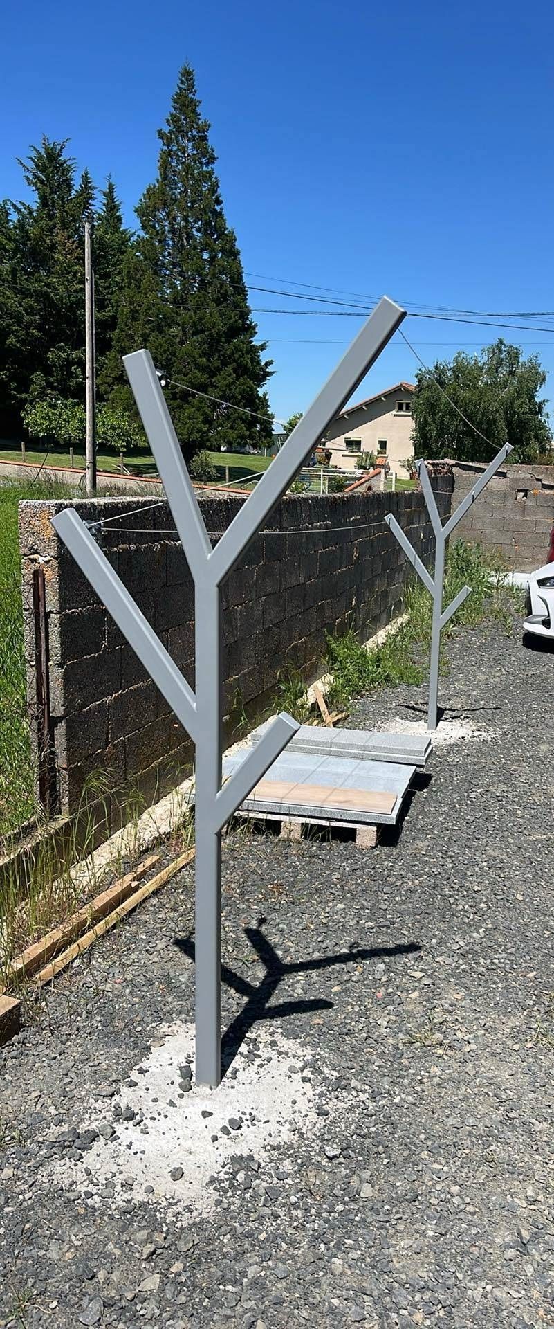Deux structures métalliques grises en forme d'arbres stylisés se dressent sur des socles en béton dans une zone extérieure gravillonnée, près d'un mur de pierre.