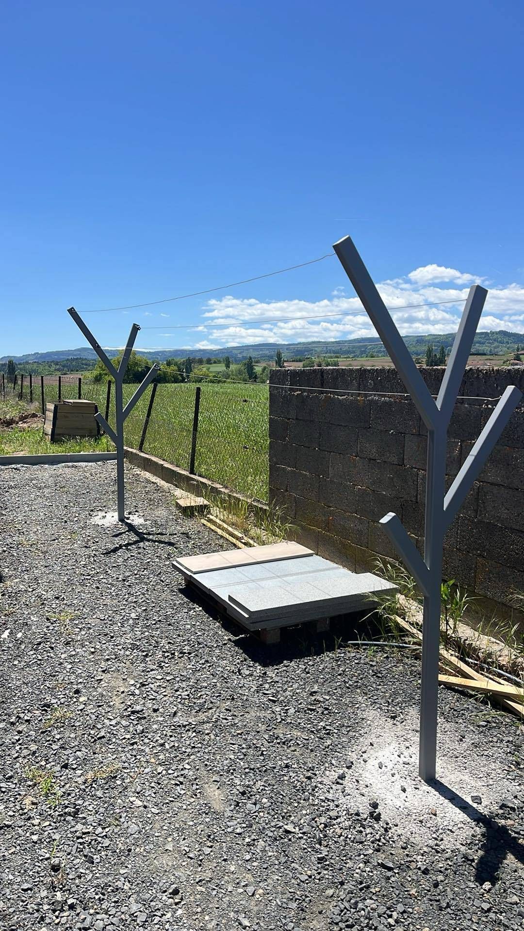 Deux hautes structures métalliques grises en forme de branches se dressent sur un sol de gravier sous un ciel d'un bleu éclatant, près d'un champ et d'un mur.