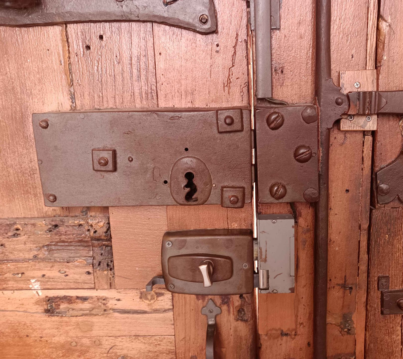 Gros plan sur une porte en bois patinée par le temps, présentant une vieille serrure à meneau en métal rouillé avec un trou de serrure et une serrure moderne à bouton rotatif grise.