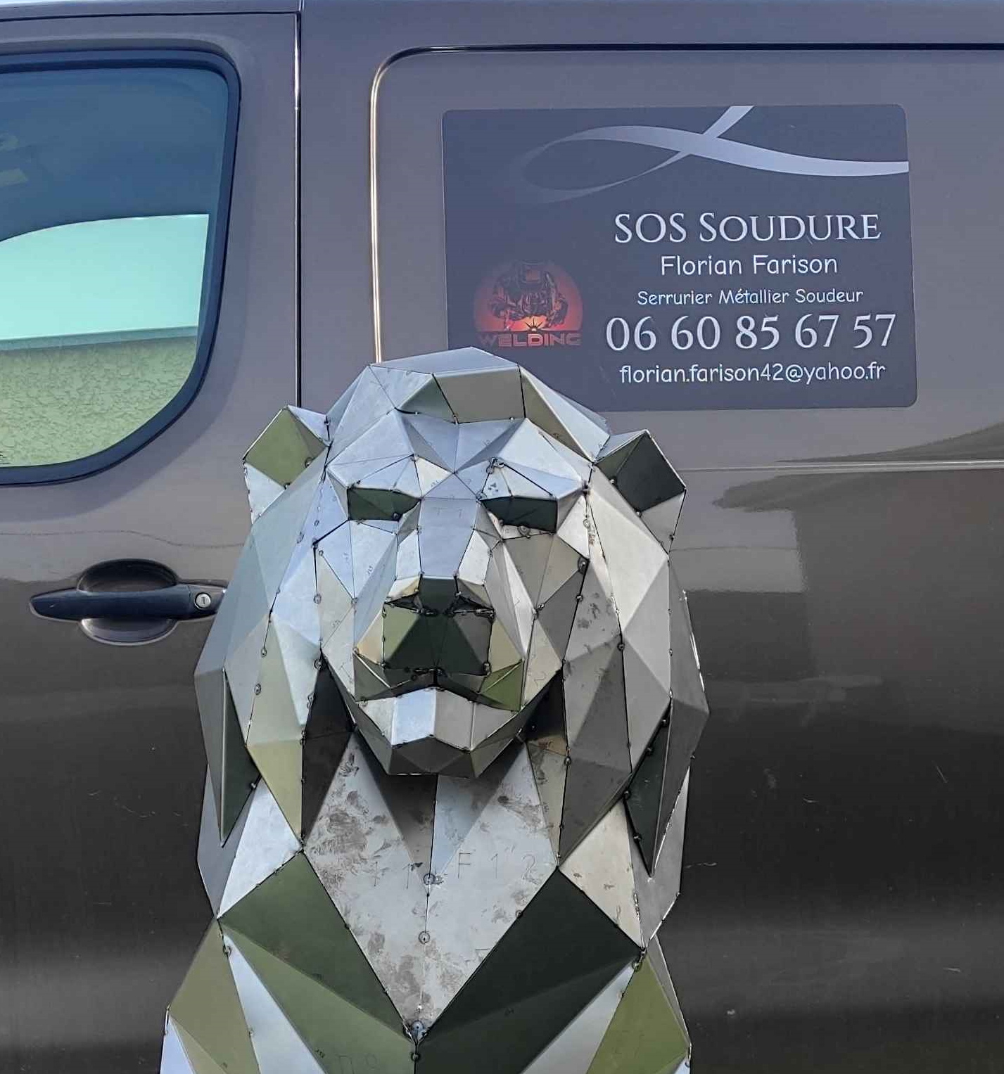 Une sculpture géométrique en métal, de style low-poly, représentant un lion dans des tons de vert olive et d'argent, posée sur un socle en béton à l'extérieur.