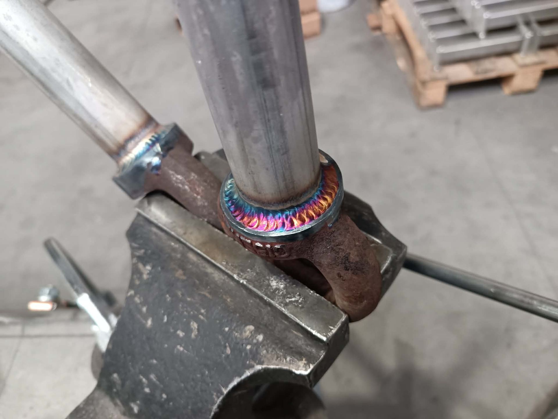 Un tuyau métallique soudé à un raccord en acier en forme de joug, fixé dans un étau métallique, laissant apparaître des cordons de soudure colorés traités thermiquement.