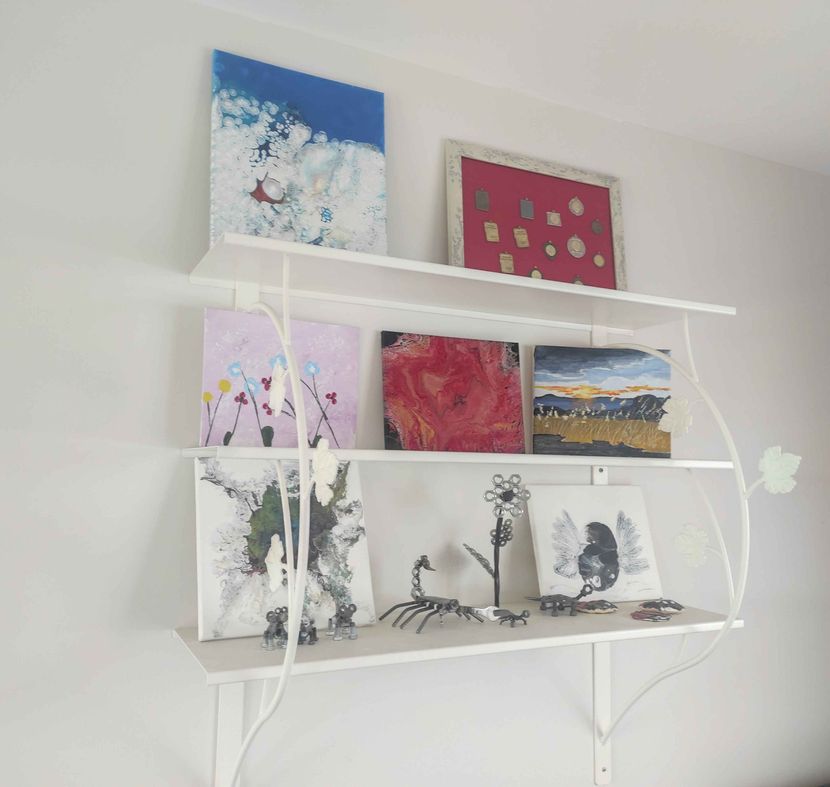 Une étagère à trois niveaux en métal blanc présente des peintures abstraites colorées, une collection d'épingles encadrées et de petites figurines en métal.