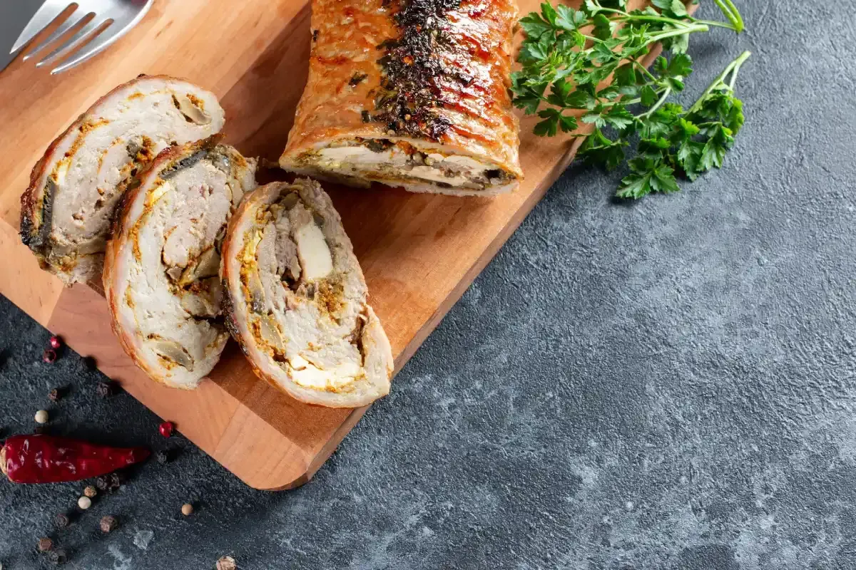 Rollito de cerdo relleno en rodajas sobre una tabla de madera con perejil y granos de pimienta.