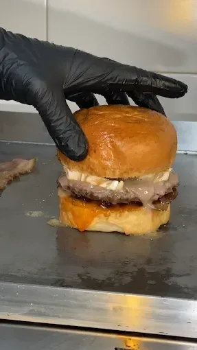 Una mano enguantada presiona una hamburguesa sobre una plancha caliente. La hamburguesa lleva dos piezas, salsa y pan.