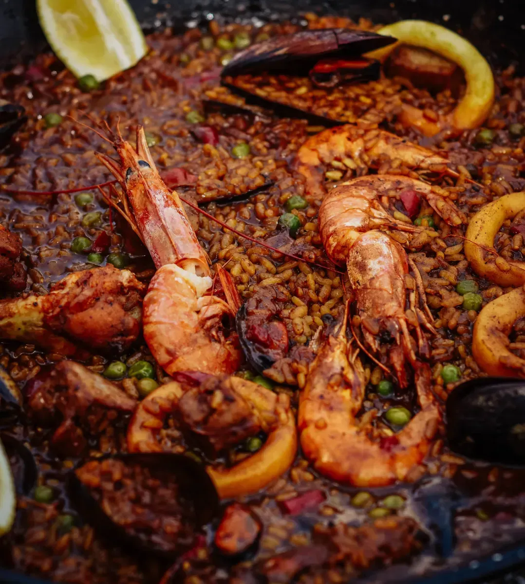 Paella con camarones, mejillones y calamares, en sartén oscura, adornada con limón.