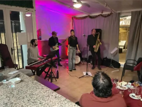 Una banda toca en una sala. Tecladista, guitarrista, cantante y saxofonista con público en mesas. Luces moradas y blancas.