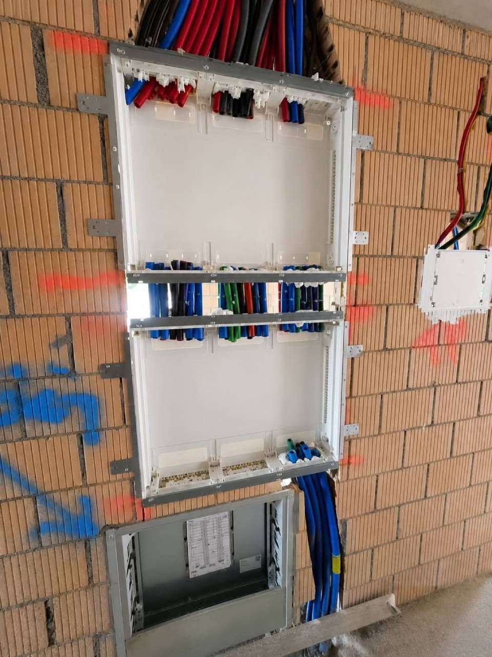 An der Wand montierte elektrische Verteilerkästen in einer Ziegelwand, mit freiliegenden Kabeln und einer unten offenen elektrischen Zugangsklappe.