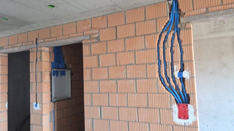 Unfertiger Backsteininnenraum mit freiliegenden blauen Elektroleitungen und einer roten Steckdose an einer Betonwand.