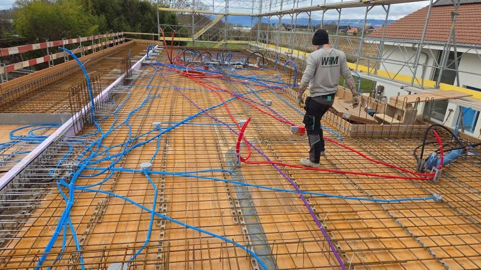 Ein Arbeiter auf einer mit Stahlbeton bedeckten Baustelle, auf deren Boden blaue und rote Kabel verlegt sind.