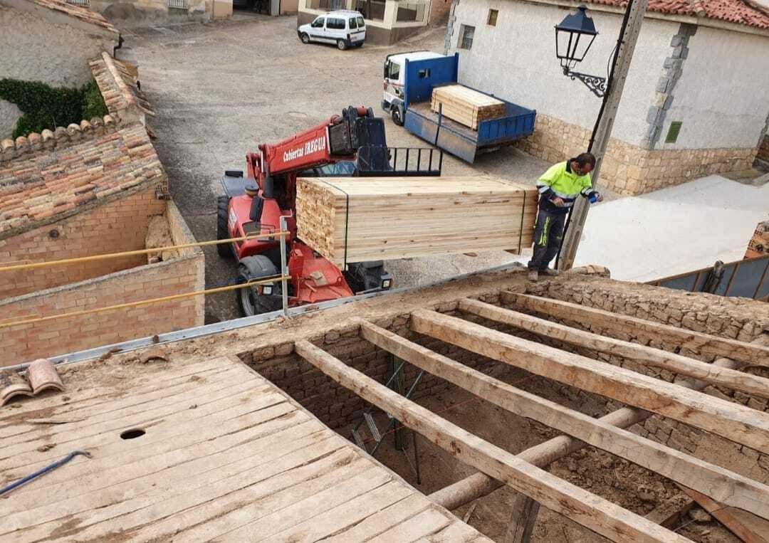 Un manipulador telescópico sube madera a un tejado para la construcción. Un trabajador con chaleco amarillo lo guía.