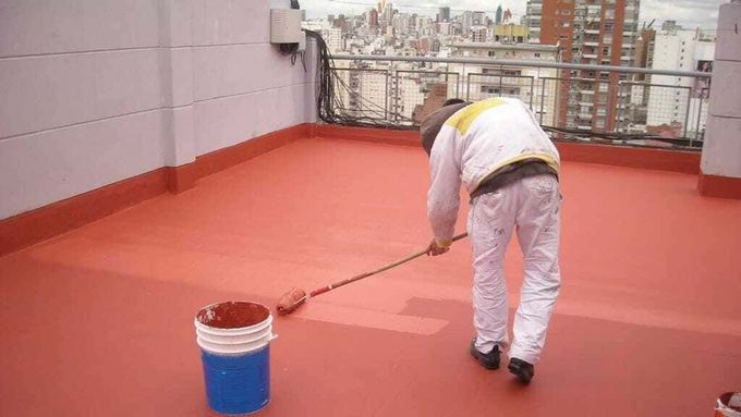 Persona pintando una capa roja sobre un tejado plano con un rodillo. Al fondo se ven edificios de la ciudad.