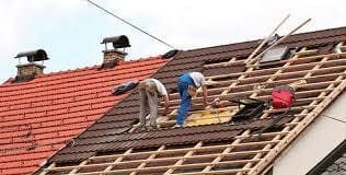 Dos techadores trabajando en el tejado de una casa, reemplazando tejas. Un lado está terminado en rojo, el otro en proceso.