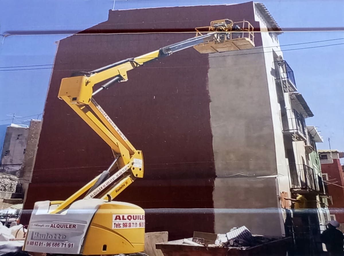 Plataforma elevadora amarilla pintando la fachada de un edificio, pintura marrón oscuro a la izquierda, color canela a la derecha, día soleado.