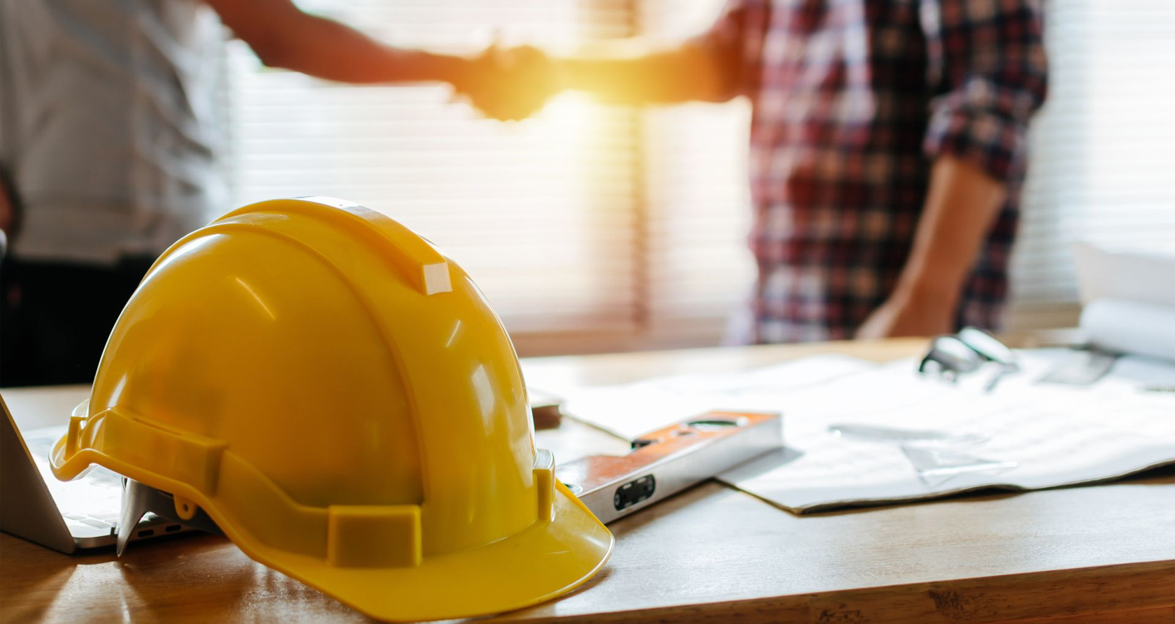 Casque jaune sur un bureau avec des plans, deux personnes se serrant la main en arrière-plan.