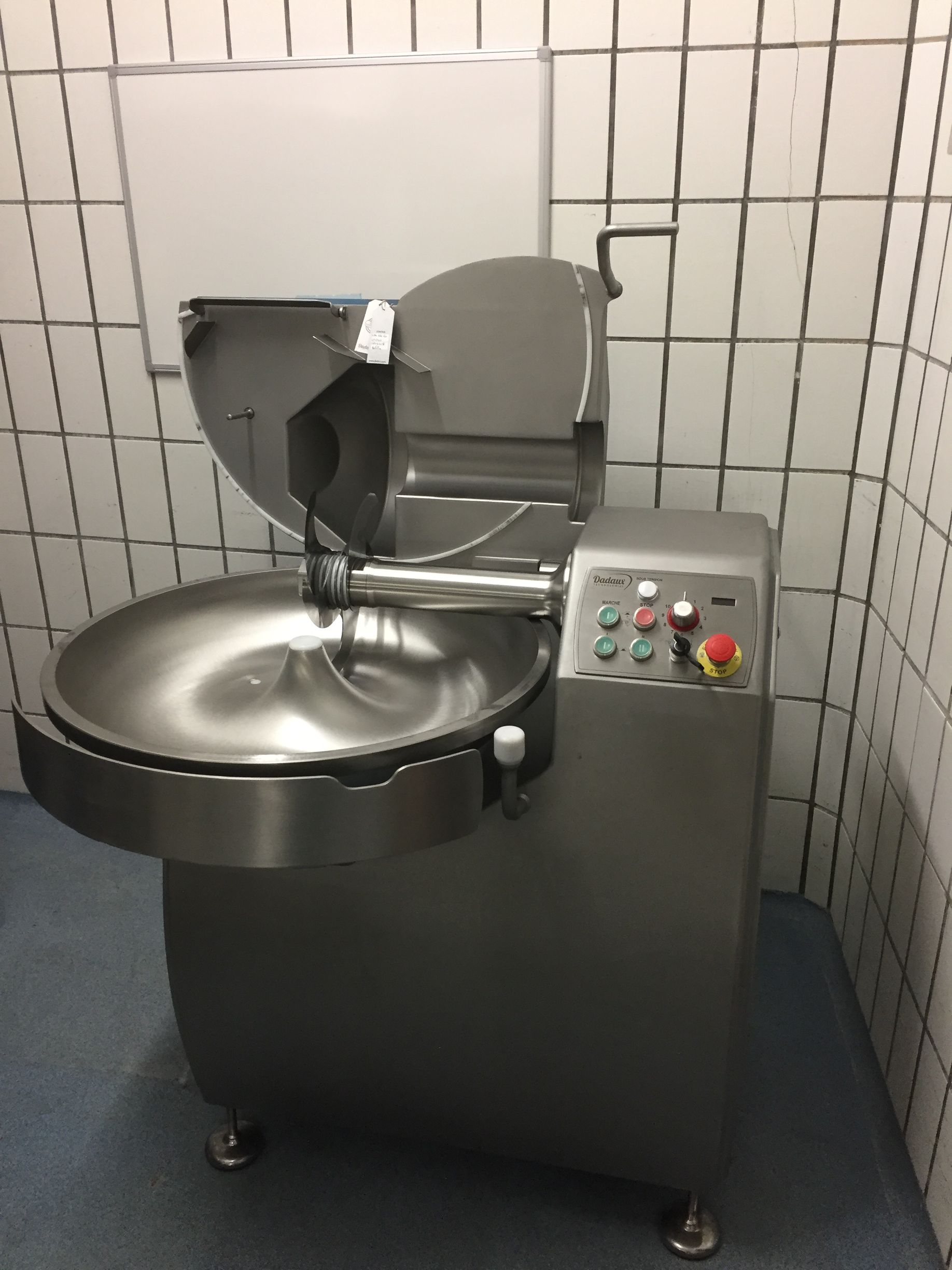 Hachoir à viande dans une pièce carrelée, une cuve et des lames en acier inoxydable, un panneau de commande avec des boutons.