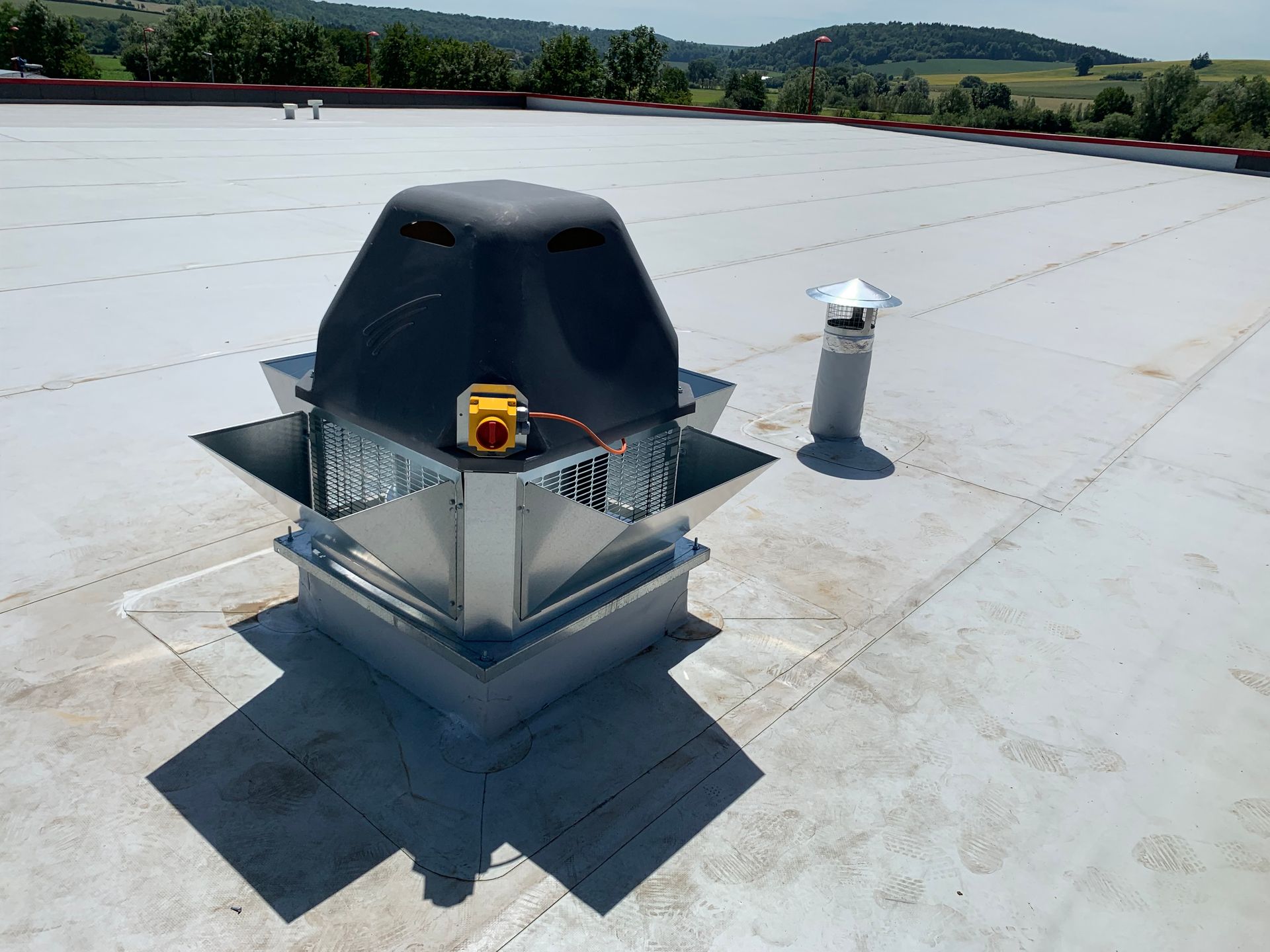 Unité de ventilation de toit avec un couvercle foncé et un interrupteur jaune, sur un toit blanc, entourée de collines verdoyantes.