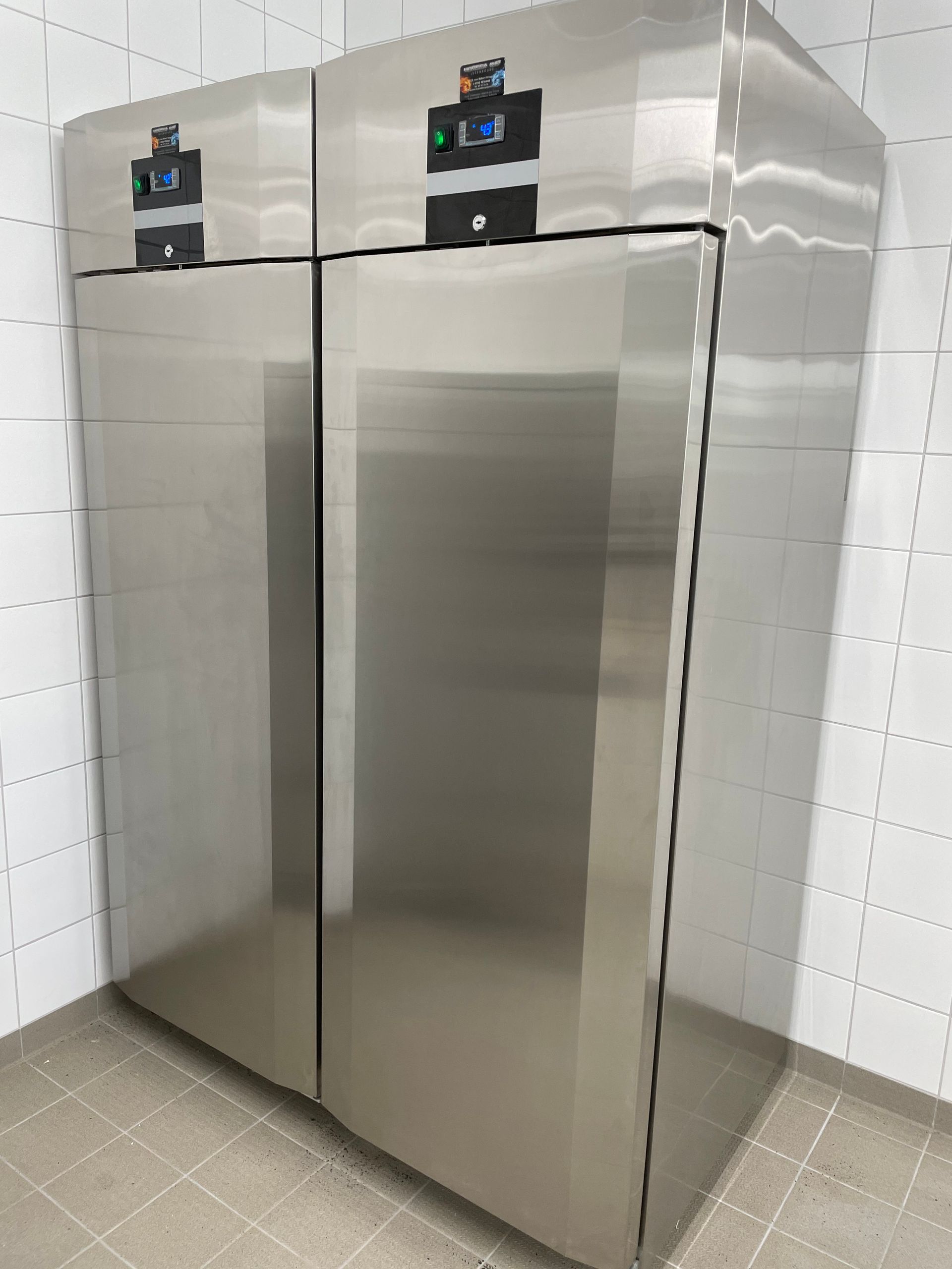 Deux grands réfrigérateurs en acier inoxydable sont placés côte à côte contre un mur carrelé.
