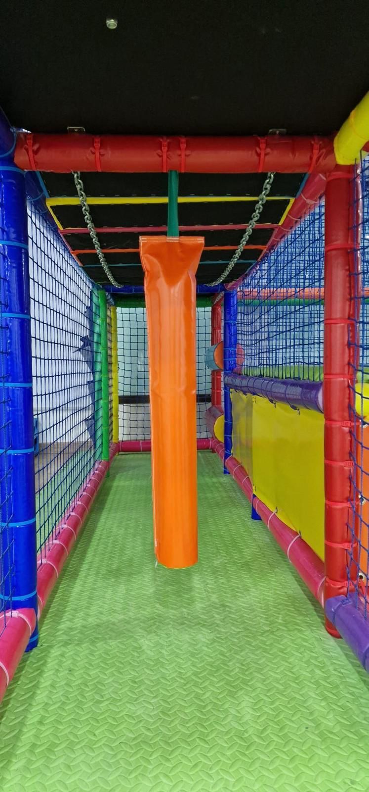 Un túnel de juegos interior con suelo verde, red de seguridad y un obstáculo colgante de espuma naranja.