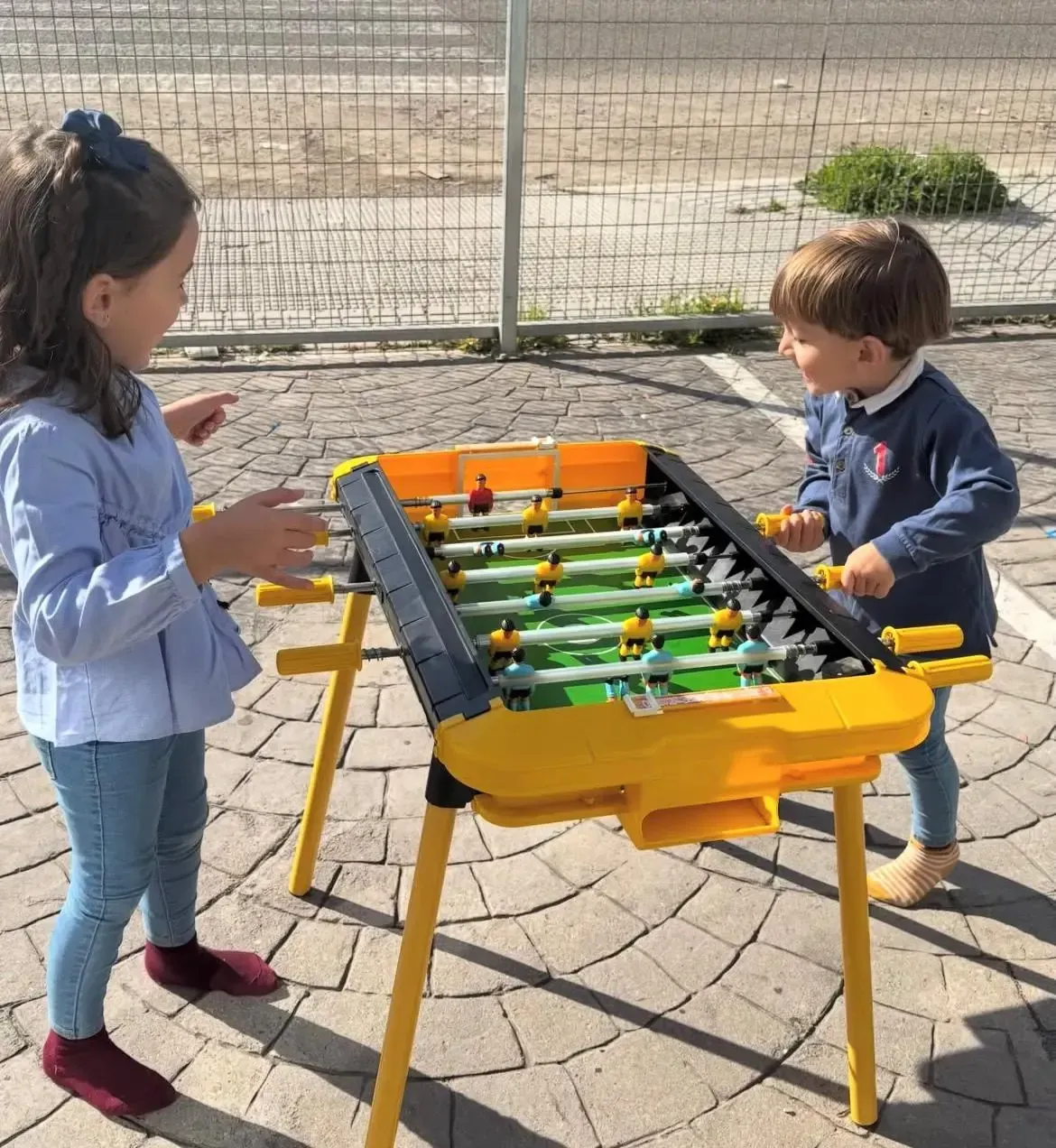 Dos niños están de pie a ambos lados de una pequeña mesa de futbolín amarilla al