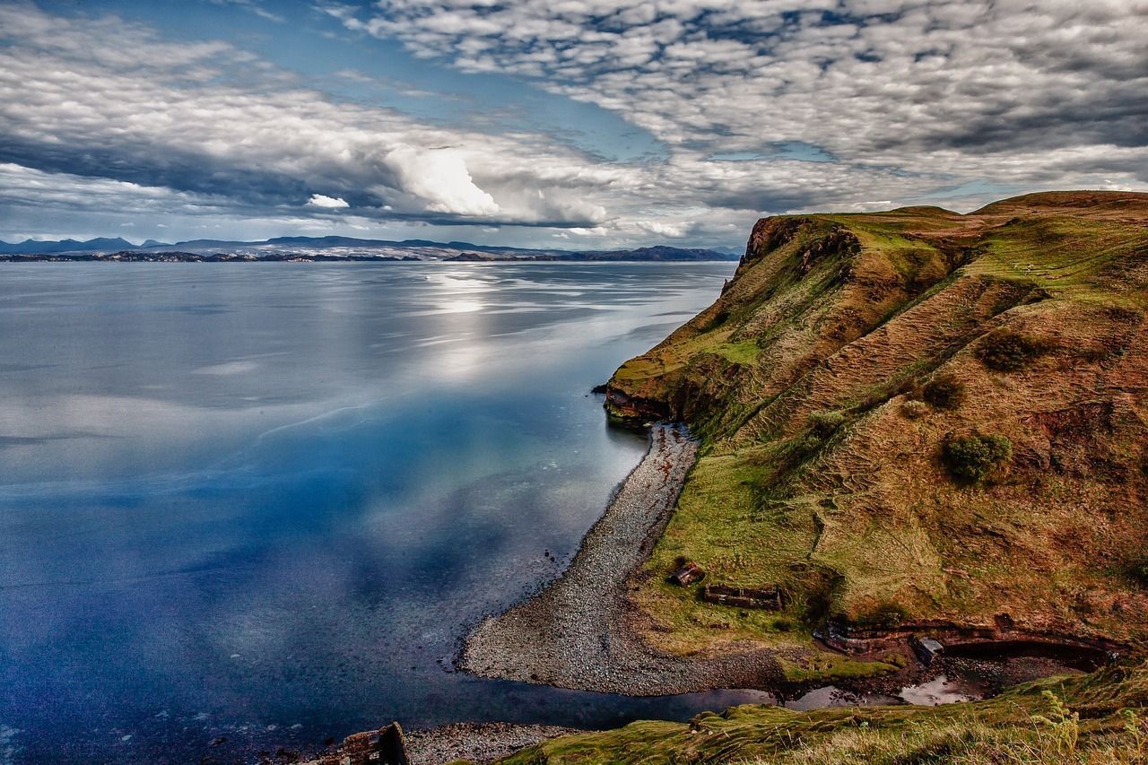 Coastal cliff and water under cloudy sky. Green and brown cliff edge, blue water, and distant land.