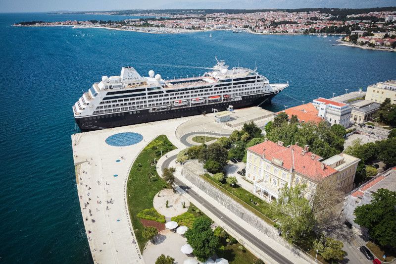 Cruise ship docked at a pier, waterfront city in the background. Blue water, clear sky.