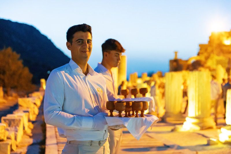 Two waiters in white uniforms serve refreshments at an ancient ruins site at dusk.