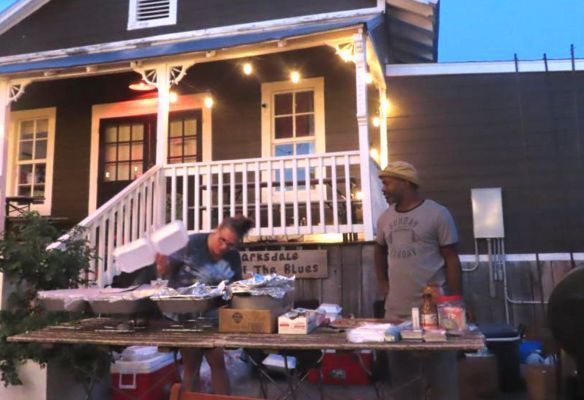 a group of people are standing around a table in front of a house having a BBQ in Clarksdale, Mississipi