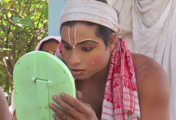 A man is looking at his face in a mirror, applying nake up for the Garamur Satra Dance