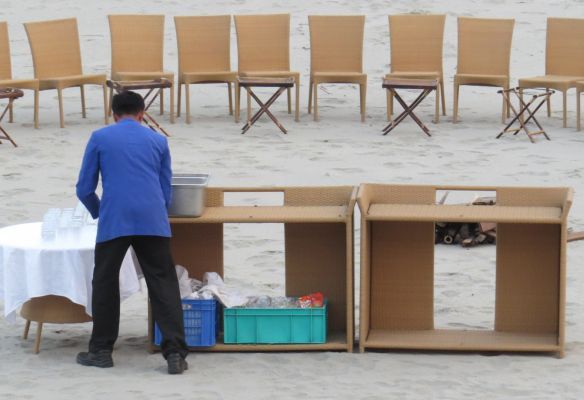 A man in a blue jacket stands in front of a table and chairs on the beach for a BBQ