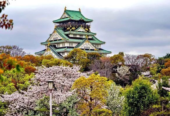 A castle with a green roof is surrounded by trees and flowers in Japan