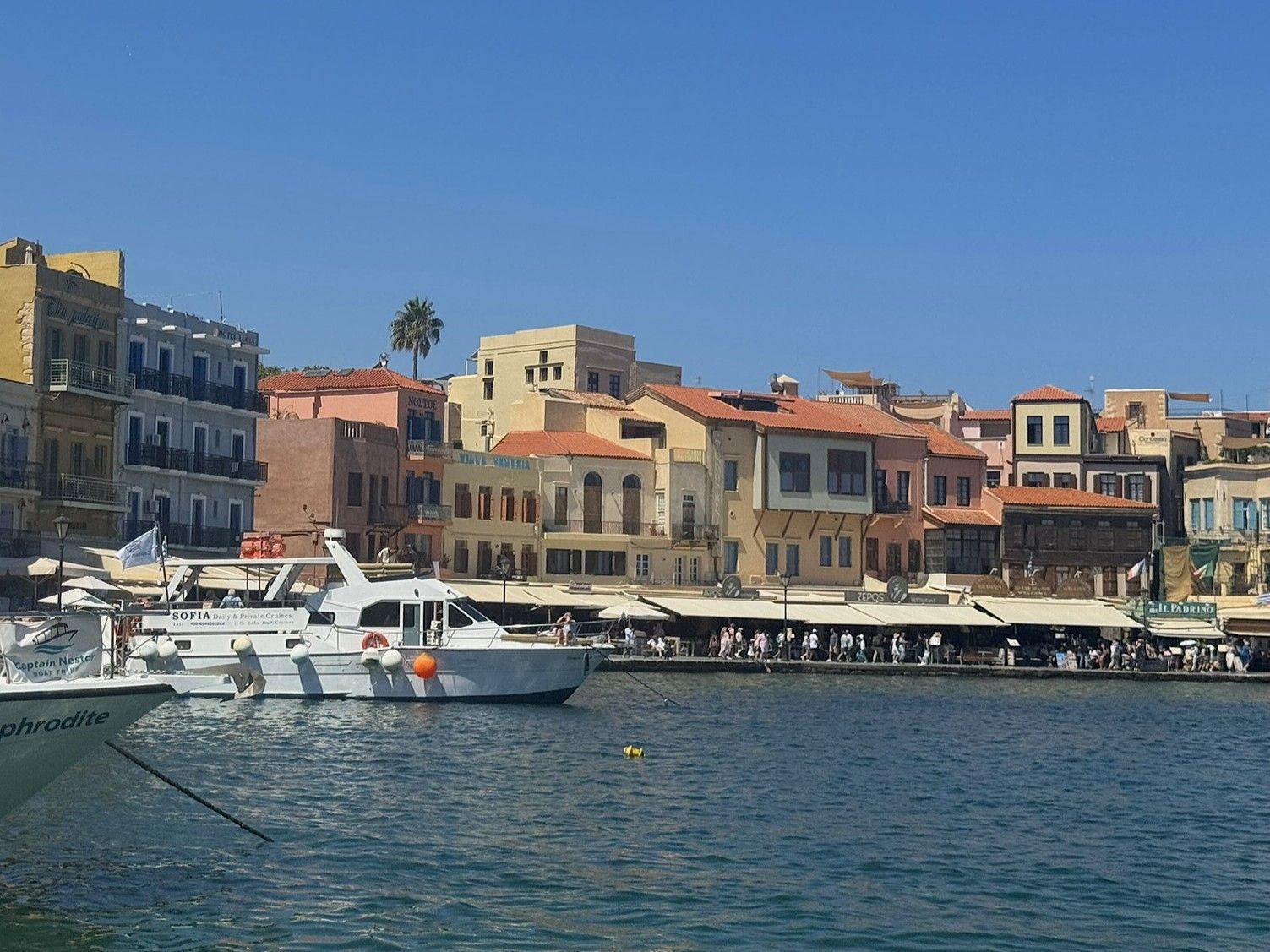 Boats on blue water, buildings with orange roofs along a harbor in Chania, Crete, Greece.