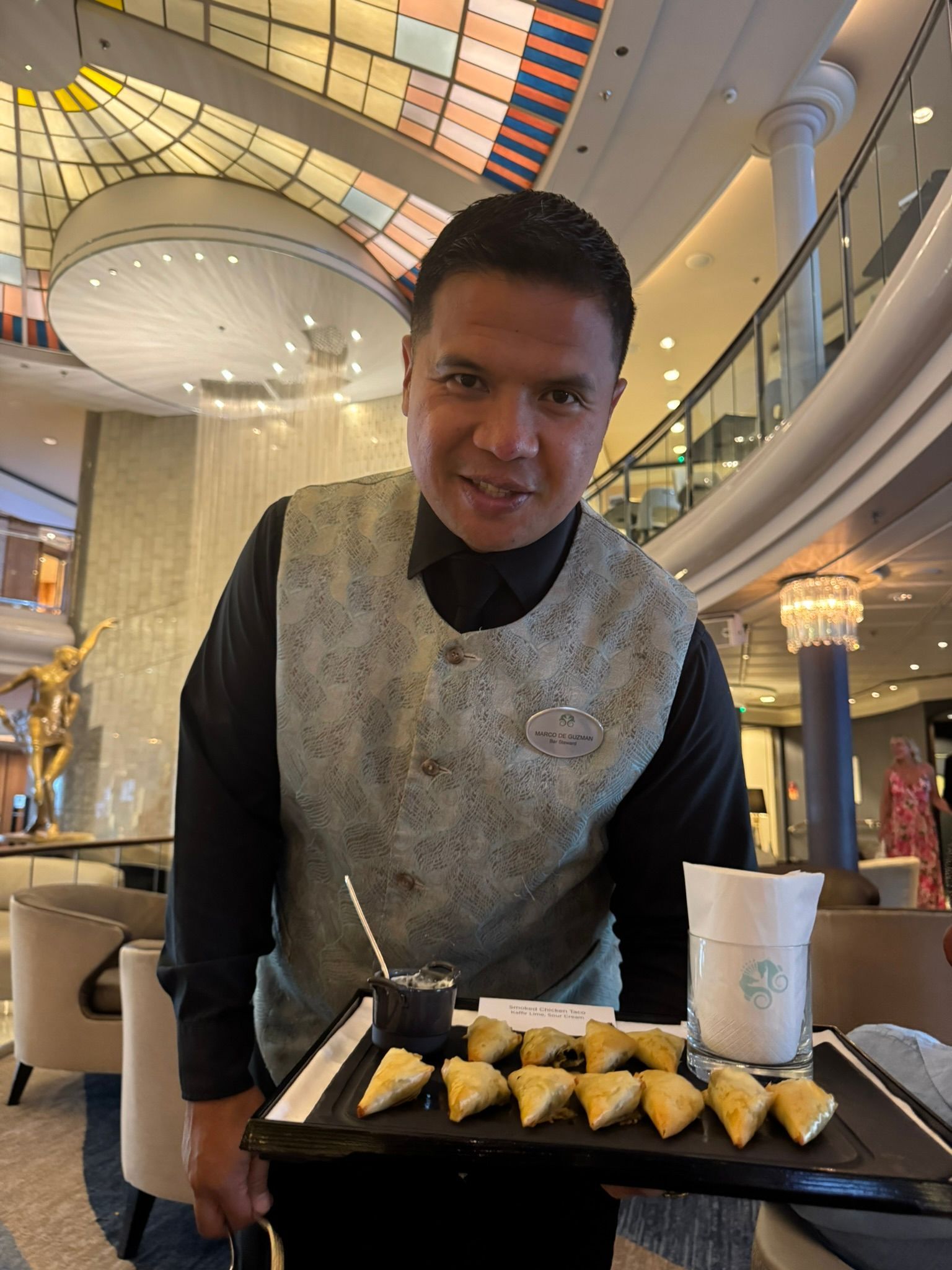 Man in vest holds tray with pastries and drink, indoors.