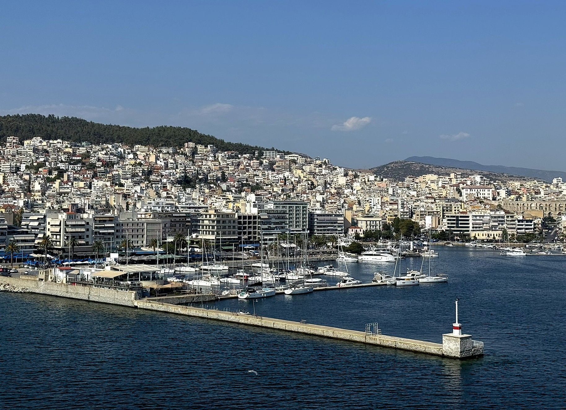 Cityscape of Kavala with boats docked at a pier, buildings clustered on a hillside, and a lighthouse.